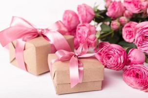 rosas frescas de color rosa pastel y cajas de regalo envueltas en papel kraft con cintas sobre una mesa de madera blanca. foto