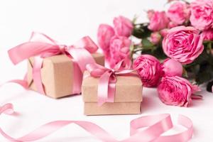 rosas frescas de color rosa pastel y cajas de regalo envueltas en papel kraft con cintas sobre una mesa de madera blanca. foto