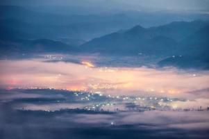 Beautiful foggy flowing on hill with illuminated city in the valley on dawn photo