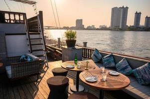restaurante en la terraza con mesa de comedor, sofá, vajilla, cubiertos en el barco a orillas del río por la noche