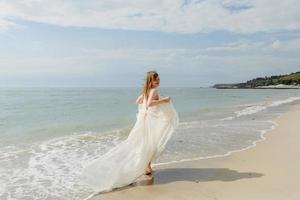 una joven hermosa con un largo vestido color leche camina por la playa y el muelle contra el fondo del mar. foto