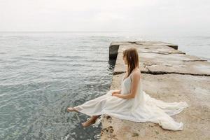 una joven hermosa con un largo vestido color leche camina por la playa y el muelle contra el fondo del mar. foto