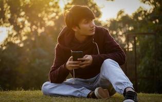 A male student using phone seated on a grass in the city park photo