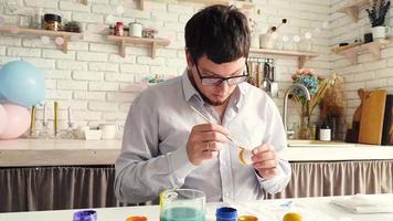 Young man coloring eggs sitting at the kitchen at home video