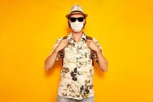 male tourist in medical mask isolated on yellow background holding his hands on straps backpack. he is dressed in Hawaiian shirt, hat, sunglasses. concept recreation, tourism, people during pandemic