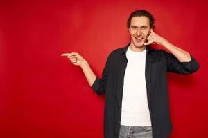 Attractive Caucasian young male in casual clothes imitates telephone conversation keeps hand near ear as if holding mobile phone has confident facial expression isolated over red background copy space photo