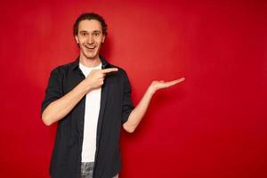 young, handsome, excited man in casual clothes, blue shirt, white T-shirt, points index finger towards hand free empty space for advertising text object isolated red background copy space. sale, promo