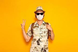 young male tourist traveler with backpack in a hat, sunglasses, a medical mask shows a peace sign with his hand isolated on a yellow background. the concept is recreation, people, adventure, vacation. photo