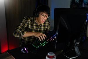 Involucró a un jugador asiático de ciberdeporte concentrado jugando videojuegos en la computadora en la habitación oscura de la noche en casa, concepto de deporte y tecnología foto