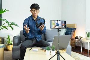 Asian man technology blogger or Social media influencer presenting and review on product by smartphone or camera on tripod recording live video for his channel on foreground creator vlogger filming. photo