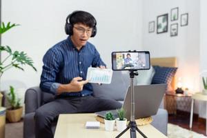 Asian man online influencer recording video live streaming, using digital smartphone camera present product review for theme about video blogging focus on camera screen show on social media.
