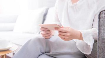 Side view of asia young woman in white sitting on sofa and using smart phone in bright living room, copy space, close up, blank for design concept photo