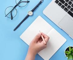 Girl write on open white book or accounting on a minimal clean light blue desk with laptop and accessories, copy space, flat lay, top view, mock up photo