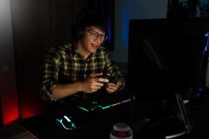 un apuesto y emocionado jugador asiático con auriculares disfruta y se regocija mientras juega videojuegos en una computadora en una habitación acogedora iluminada con luz cálida y de neón, juegos y tecnología concepto de deporte electrónico foto