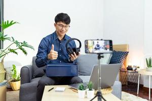 Asian man technology blogger or Social media influencer presenting and review on product by smartphone or camera on tripod recording live video for his channel on foreground creator vlogger filming. photo
