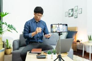 Young asian male blogger recording vlog video on camera review of product at home office, Focus on tripod mounted camera screen broadcast live stream video to a social network. photo