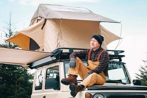 Adventurer man sitting down on roof of the car with tent mounted on it for free travel camping lifestyle