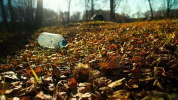 garrafa de plástico transparente grande vazia entre folhas caídas marrons contra árvores no ambiente poluente do parque closeup câmera lenta video