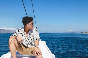 Asian man wearing sunglasses and wireless earphone sitting on a boat while looking to the sea photo