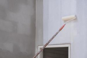 cepillo de rodillo de mango largo que aplica pintura blanca de imprimación con marco de puerta en la pared de cemento dentro del sitio de construcción de la casa, concepto de renovación de la construcción y la casa foto