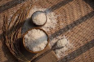 Sunlight and shadow on surface of white rice in ceramic bowl and wooden spoon with ear of paddy on threshing basket background, top view with copy space photo