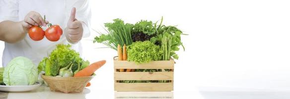 pancarta de fondo de alimentos saludables de una mujer con sobrepeso sosteniendo tomates frescos y mostrando el pulgar hacia arriba con varias verduras orgánicas en una cesta de bambú y una caja de madera en una mesa de mármol en una cocina blanca foto