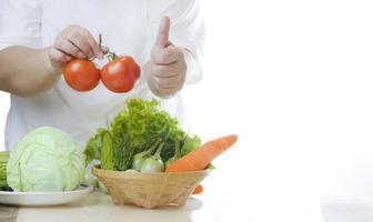 Cropped image of fat woman holding fresh tomatoes and showing thumps up with non-toxic vegetables in a bamboo basket on marble table in white kitchen, healthy food for overweight people concept photo