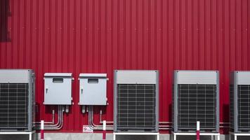 Row of air conditioner compressors with 2 electric control cabinets and electric pipelines system on red metal sheet wall outside of building photo