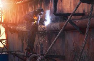 Side view of welder in protective workwear on platform is welding the old rusty vessel hull in shipyard area at sunset time photo