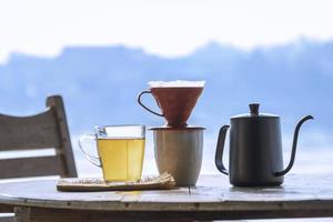 Glass cup of hot aromatic tea with drip coffee cup set on wooden round table against riverside view background at morning time photo