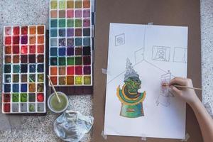 Top view of young girl's hand is painting art picture on paper with 2 watercolor trays and artistic things on white marble table photo