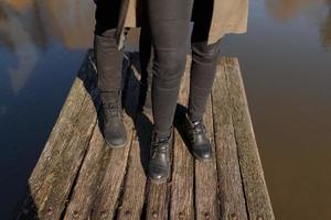 Cropped photo young legs in sneakers, married couple, husband and wife on a wooden bridge near lake. Rear view of couple standing on pier. lower half. Place for text and design. Summer in love.