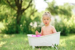 Happy toddler girl takes a milk bath with petals. Little girl in a milk bath on a green background. Bouquets of pink peonies. Baby bathing. Hygiene and care for young children. photo