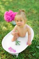 Happy toddler girl takes a milk bath with petals. Little girl in a milk bath on a green background. Bouquets of pink peonies. Baby bathing. Hygiene and care for young children. photo
