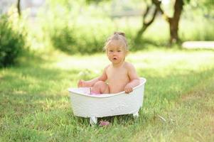 Happy toddler girl takes a milk bath with petals. Little girl in a milk bath on a green background. Bouquets of pink peonies. Baby bathing. Hygiene and care for young children. photo