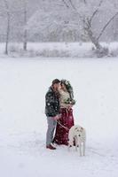 Beautiful bride and groom with a white dog are standing on the background of a snowy forest. photo