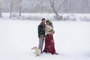 Beautiful bride and groom with a white dog are standing on the background of a snowy forest. photo