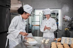 dos chefs profesionales con uniformes y delantales de cocina blanca amasan masa de pastelería y huevos, preparan pan, galletas y comida fresca de panadería, horneando en horno en una cocina de acero inoxidable de un restaurante. foto