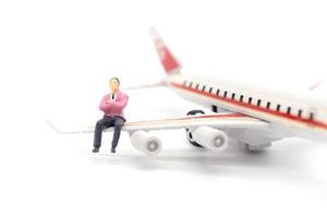 Empresario en miniatura sentado en el ala del avión sobre fondo blanco.