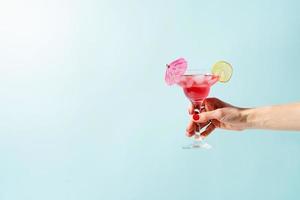 female hand with nice red manicure holding fresh summer cocktail with strawberry, lime and ice cubes on blue background with copy space photo