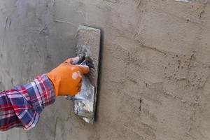 las manos de un trabajador que se está enyesando de cerca llevan guantes de goma naranja para evitar que el cemento les muerda las manos, construyendo la pared de la casa foto