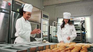 Two professional female chefs in white cook uniforms and aprons knead pastry dough and eggs, prepare bread, cookies, and fresh bakery food, baking in oven at a stainless steel kitchen of a restaurant. video