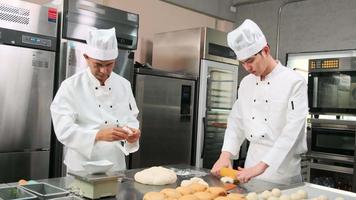 zwei professionelle asiatische männliche köche in weißen kochuniformen und schürzen kneten teig und eier, bereiten brot und frische backwaren zu und backen im ofen in der edelstahlküche des restaurants. video