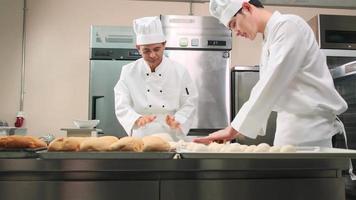 due cuochi professionisti asiatici maschi in uniformi bianche da cuoco e grembiuli stanno impastando pasta e uova, preparando pane e prodotti da forno freschi, cuocendo in forno nella cucina in acciaio inossidabile del ristorante. video