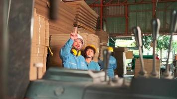 ingegnere maschio asiatico in uniforme di sicurezza e elmetto e collega lavoratrice ispeziona lo stoccaggio, l'ordine di scorta nel magazzino di fabbrica, le pile di cartone impilabile, la gestione del prodotto industriale. video