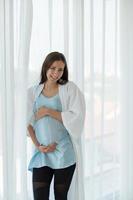 A pregnant woman in a bedroom with white curtains and early morning atmosphere photo