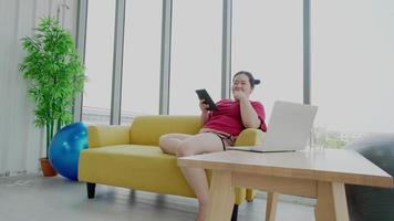 Overweight or fat young woman looking on laptop she sitting in living room and eat crisps and drink water at home. She having fun happy and smile. video