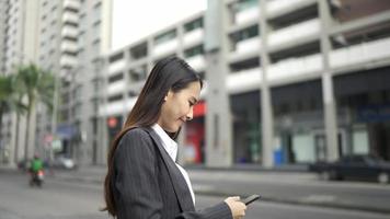 mujer asiática sonriente con traje formal usando teléfono móvil escribiendo mensajes de texto caminando sosteniendo una computadora portátil fuera de negocios teléfono celular ciudad smartphone cámara lenta video