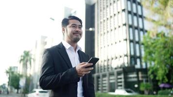 empresário asiático sorrindo vestindo um terno preto está andando nas ruas da cidade para trabalhar. na mão segurando usando um smartphone. conceito de estilo de vida urbano. video