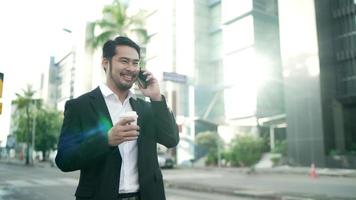 un hombre de negocios asiático sonriendo con un traje negro camina por las calles de la ciudad para trabajar. en la mano sosteniendo una taza de café usando un teléfono inteligente. concepto de estilo de vida urbano.
